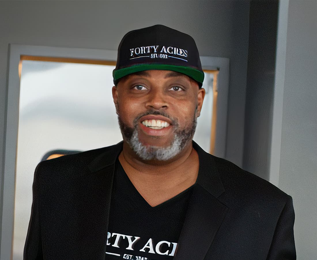A man in a black hat and shirt that say 'Forty Acres' standing in front of a kitchen door in a restaurant