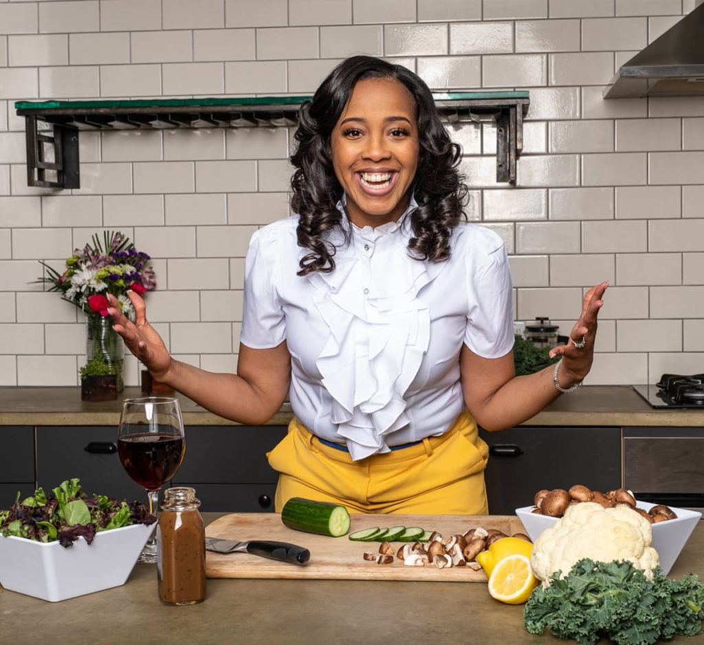 A woman standing in front of a chopping board in a kitchen with a salad and wine on the counter