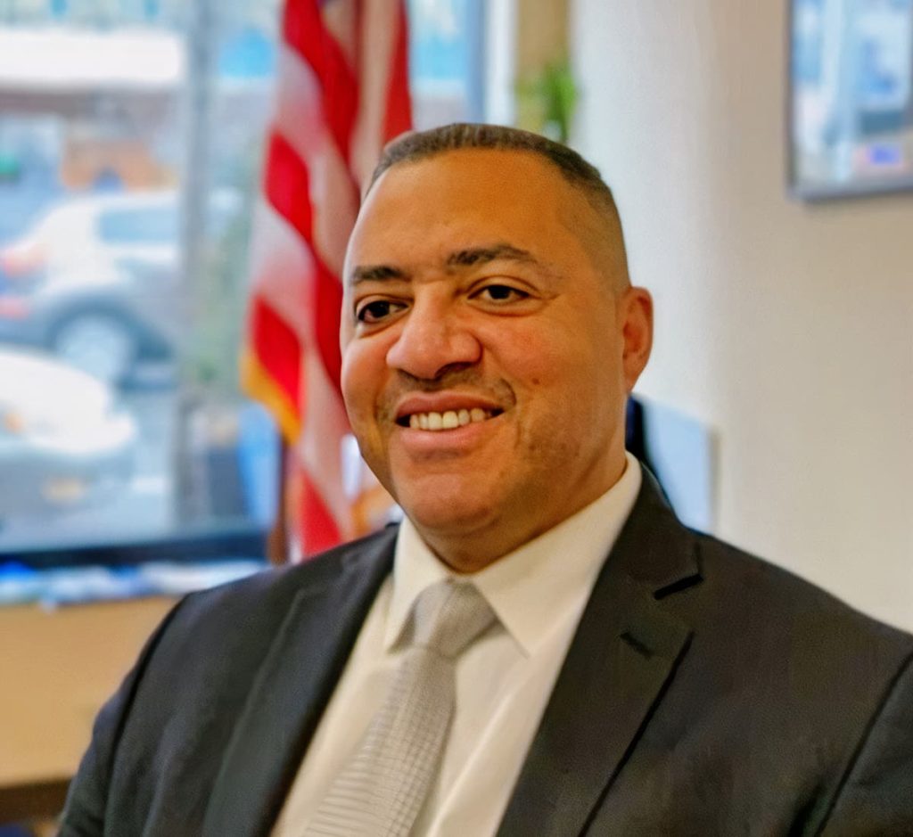 A latino man in a suit sitting at a desk in front of the American flag