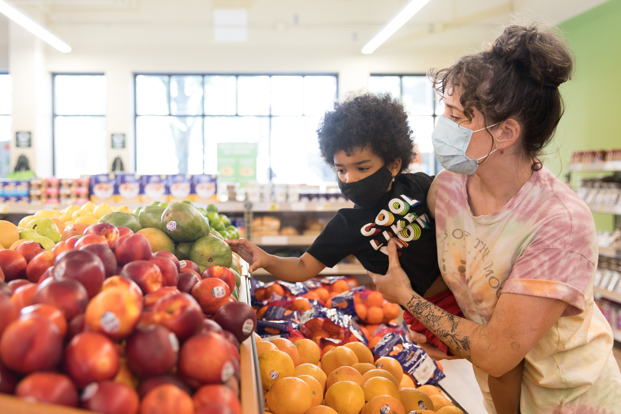 A woman wearing a face mask is holding a young boy who is selecting a mango from a grocery shelf