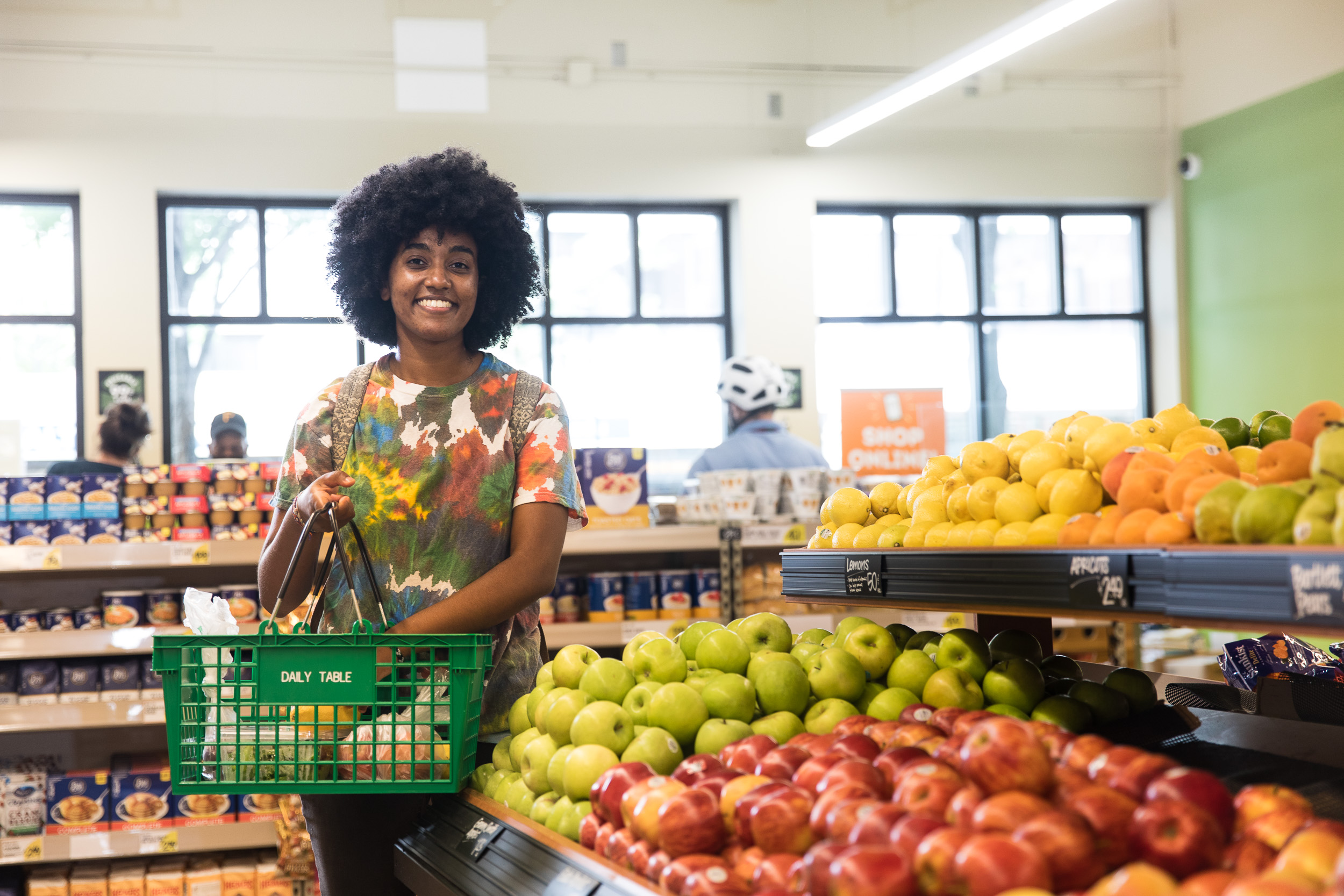 A Black woman with a colourful shirt is smiling at the camera while standing amongst produce at a grocery store