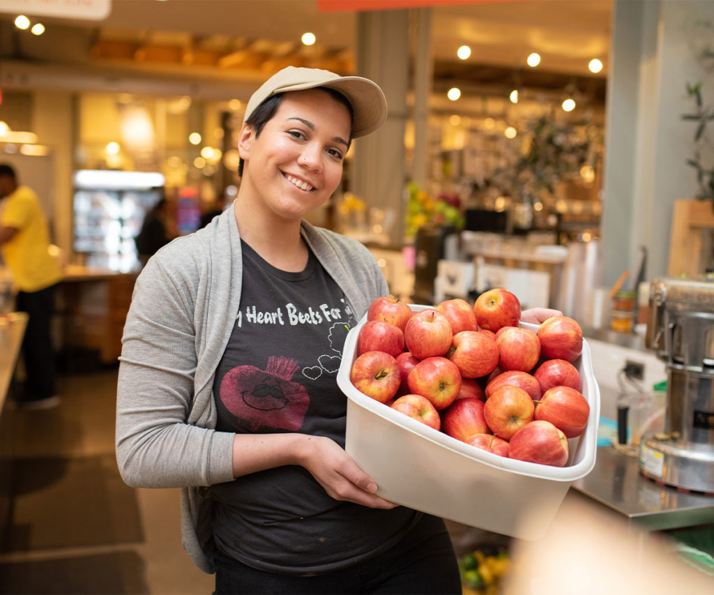 A woman with short hair and a beige hat is smiling while holding a basket of red apples