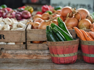 Fresh zucchini, carrots, onions, and garlic displayed in baskets and crates at a farmers market.