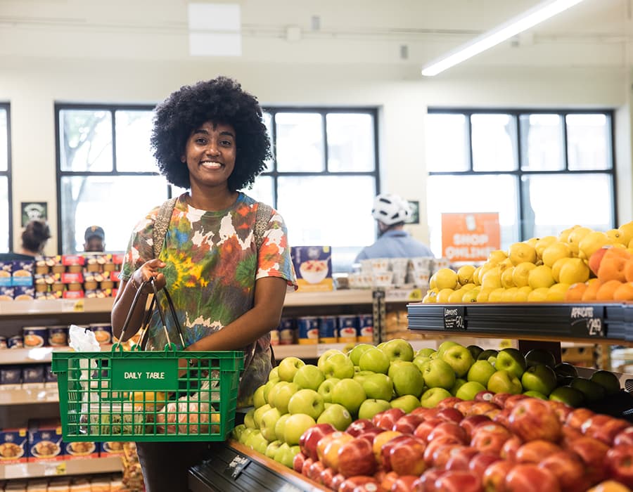 A woman in a tie-die shirt is standing in a produce aisle and adding apples to a green basket