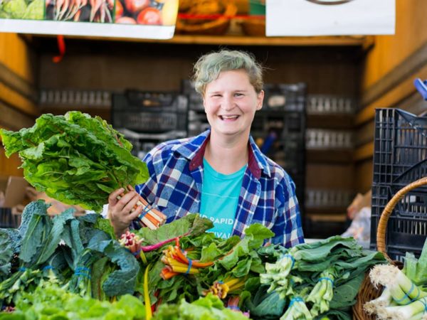A person standing behind a farm stand full of collard greens while smiling