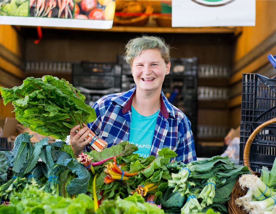 A person standing behind a farm stand full of collard greens while smiling