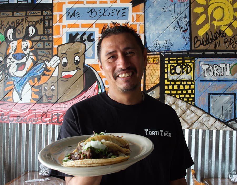 A Latino entrepreneur from Torti Taco smiling and holding out a plate of tacos in his restaurant