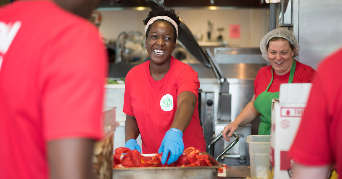 A group of four people in bright red t-shirts smiling while cooking peppers in an industrial kitchen