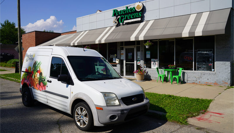 Exterior of Peaches & Greens, a neighborhood produce market in Detroit, Michigan. The store's logo sits atop the storefront with a striped awning and a few green tables and chairs are set up outside. As compact van is packed out from with the Peaches & Greens logo graphic visible on the side.