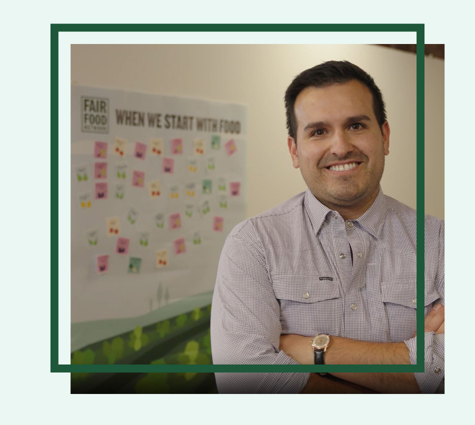 Alex Canepa, Policy Director at Fair Food Network, stands with his arms crossed in front of an office wall with a printed banner and sticky notes.