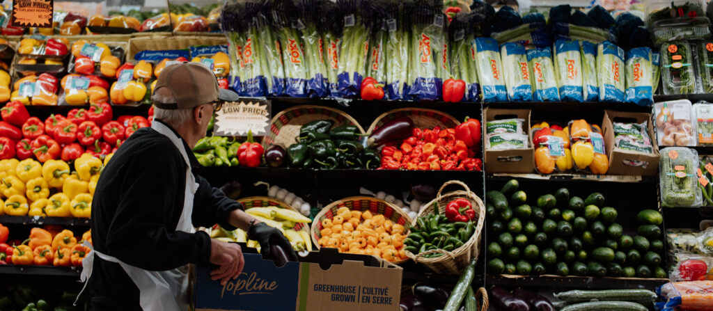 A worker stocks fresh vegetables and fruits at a grocery store produce section, surrounded by colorful produce.
