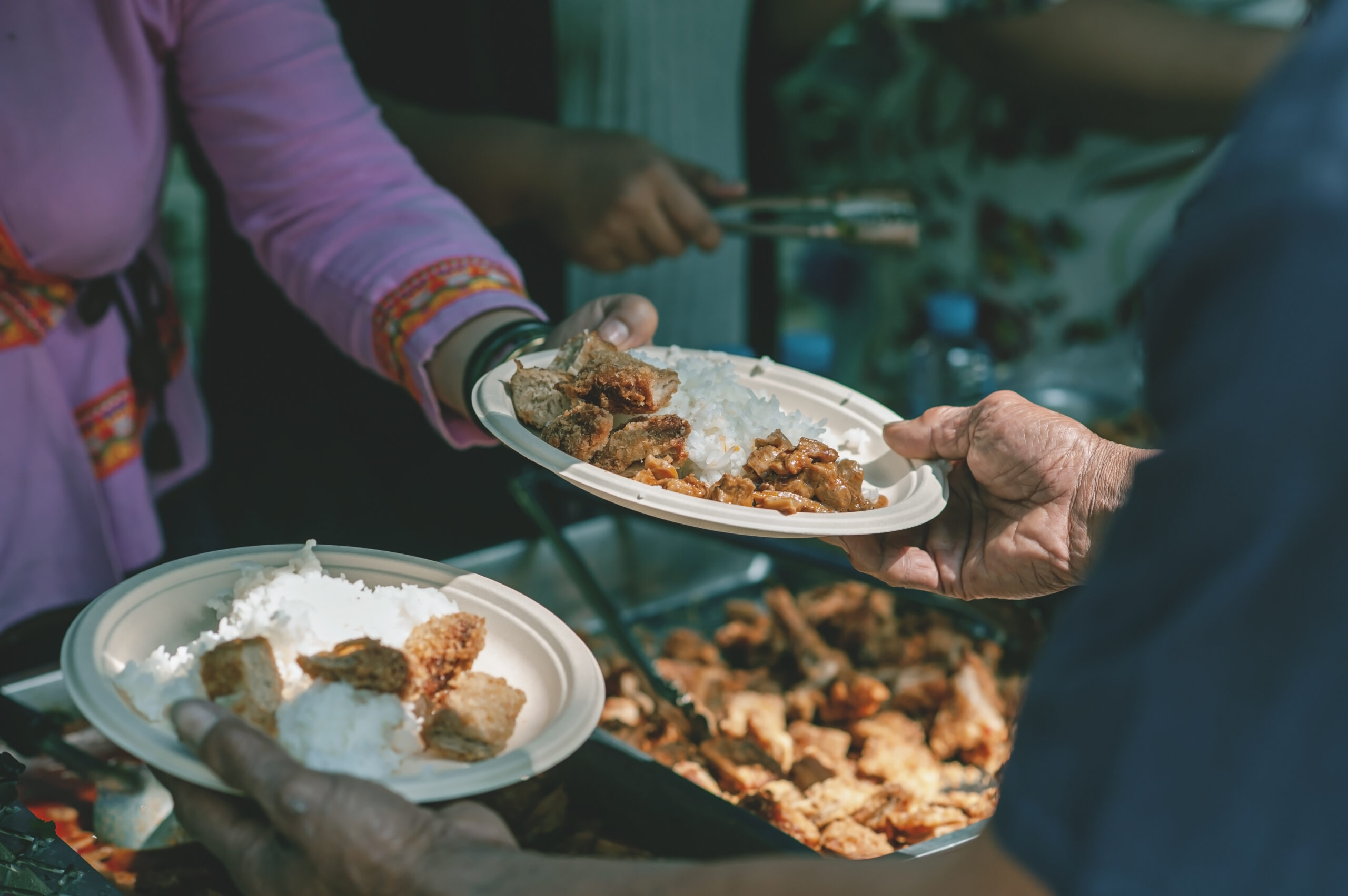 Hand offering plate of white rice and fried meat to another hand with a backdrop of other similar plates and vats of rice and meat.