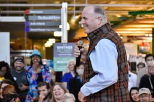 A man in a plaid vest speaks into a microphone to a smiling crowd inside a public building.