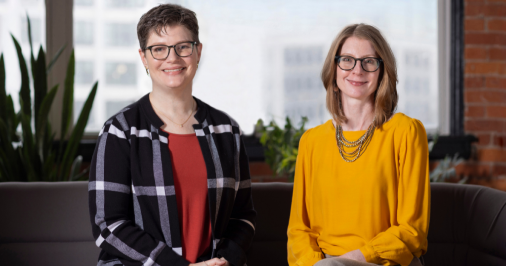 Two women sit indoors on a couch, smiling at the camera, with plants and windows in the background.