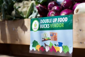 Sign for Double Up Food Bucks Vendor in front of fresh onions and leafy produce at a market stand.