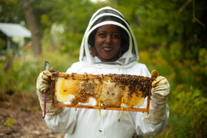 A beekeeper in protective gear holds a honeycomb frame covered with bees outdoors.