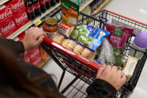 A person pushes a grocery cart filled with various food items and a bottle of water in a store aisle.