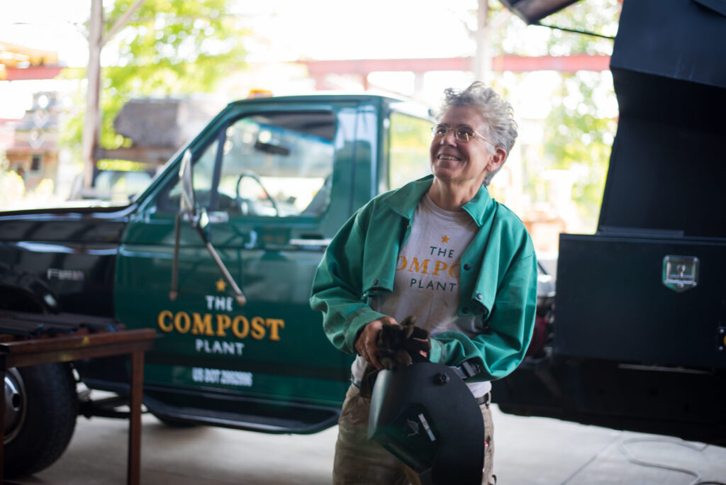 Smiling person in green jacket holds welding helmet near a green truck labeled The Compost Plant..