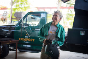 Smiling person in green jacket holds welding helmet near a green truck labeled The Compost Plant..