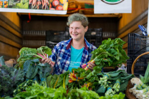 Person smiling behind a market stall, holding fresh leafy greens among assorted vegetables.