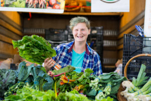 Smiling person stands behind a stall with leafy greens and vegetables at a market.