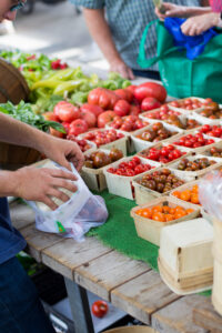 A person bags produce at a market stall with tomatoes and vegetables in small baskets.