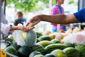 A person pays with coins for a watermelon at an outdoor market stand.