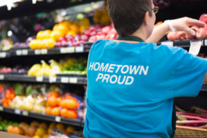Person in a HOMETOWN PROUD shirt arranges produce in a grocery stores fruit and vegetable section.