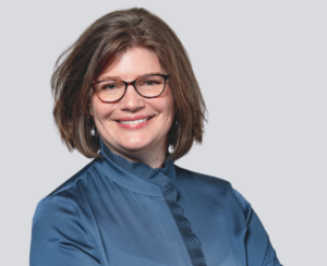 Woman with short brown hair and glasses, smiling, wearing a blue blouse, on a light gray background.