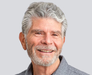 Smiling older man with gray hair and beard wearing a gray collared shirt against a light background.