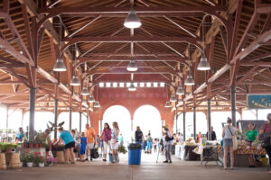 People shopping at an open-air market under a large pavilion with tables of goods and high wooden beams.