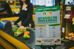 Sign about double food bucks for fruits and veggies at a grocery store, with produce on the checkout.