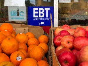 Oranges and apples in boxes at a market with signs for EBT and WIC benefits in the background.