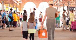 A woman and two children walk through a busy outdoor market, carrying a bright orange tote bag.