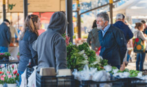 A man and two women talk at a busy outdoor farmers market produce stand on a sunny day.
