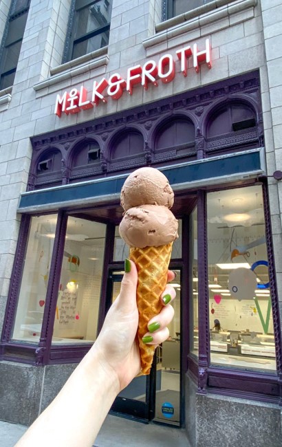 A hand with green nails holds a double-scoop chocolate ice cream in front of Milk & Froth shop.