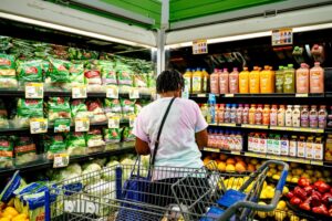 Person shopping in a grocery store produce section, facing shelves of salad bags and refrigerated drinks.