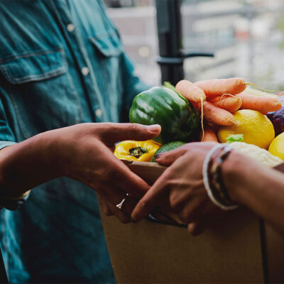 Two people exchange a box filled with fresh vegetables, including peppers, carrots, and lemons.