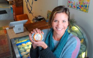 Smiling woman in teal scarf holds a cheese round in a shop with a counter and colorful decor.