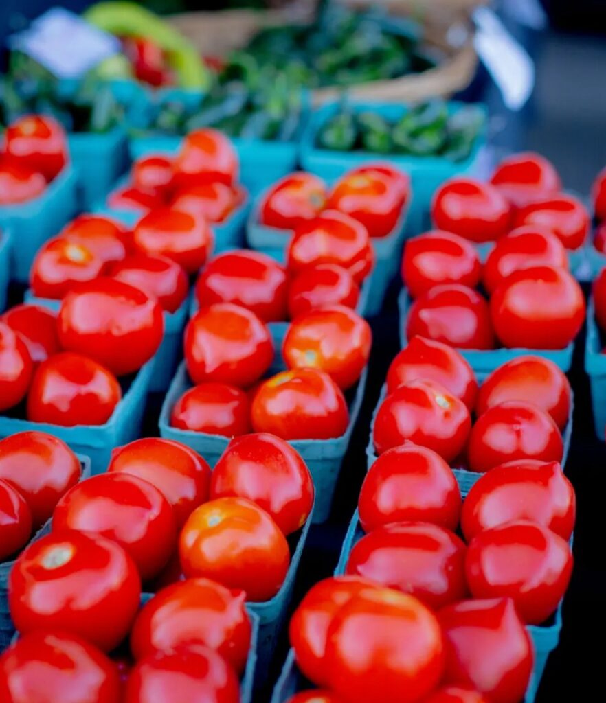 Bright red tomatoes displayed in blue cartons at a market stand, with green vegetables in the background.