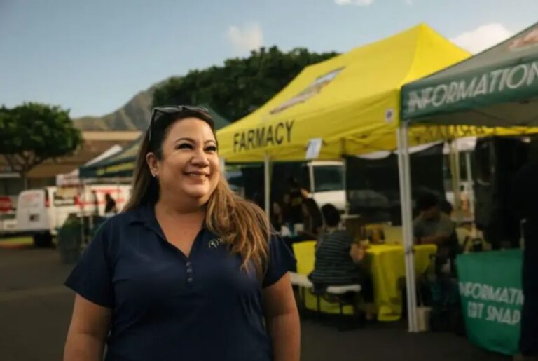 Smiling woman stands outside near yellow tents at a market event with trees and mountains in the background.