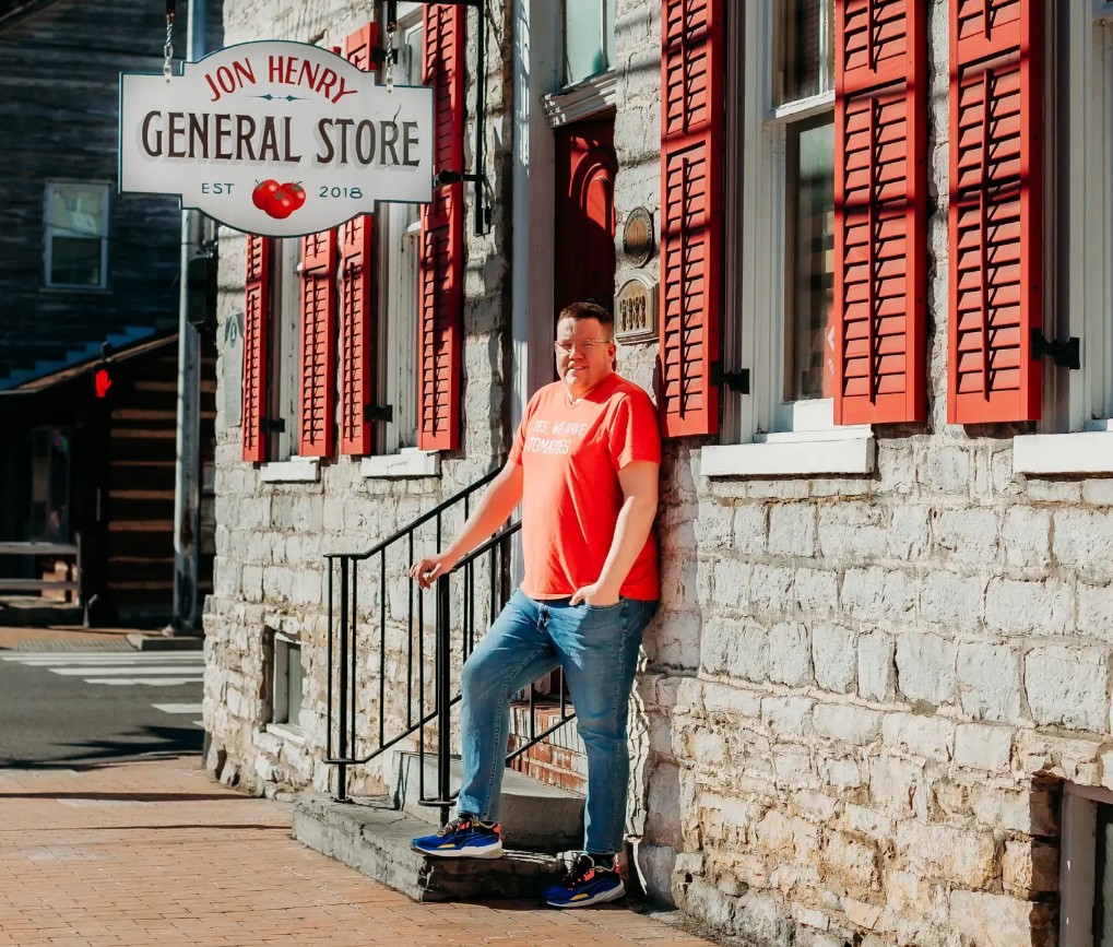 Person in a red shirt stands outside Jon Henry General Store, next to a stone building with red shutters.