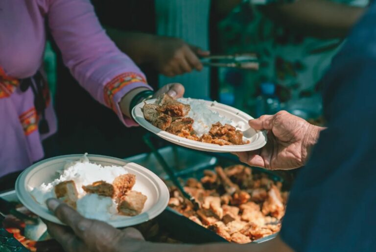 Two people hold plates of rice and fried food at an outdoor food stall or buffet.