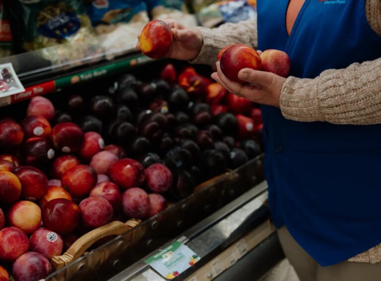 Person in a blue vest holds two nectarines while standing by a display of plums and nectarines in a store.