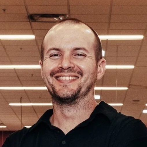 A man with a shaved head and beard, smiling, stands indoors under ceiling lights.