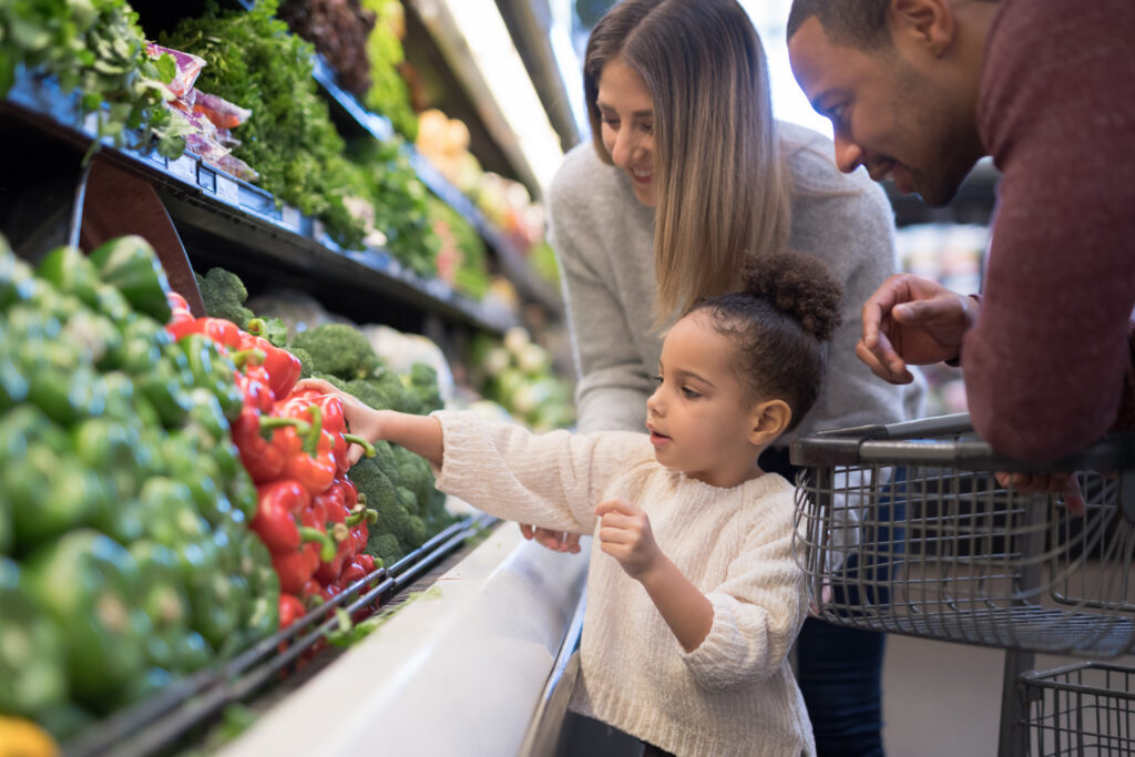 A child picks red bell peppers in a grocery store, with two adults watching and smiling nearby.