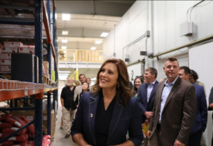 A woman in business attire smiles while touring a warehouse with a group of people behind her.