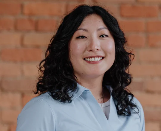 Woman with curly black hair smiling, wearing a light blue shirt, standing in front of a brick wall.