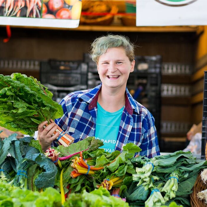 A person standing behind a farm stand full of collard greens while smiling