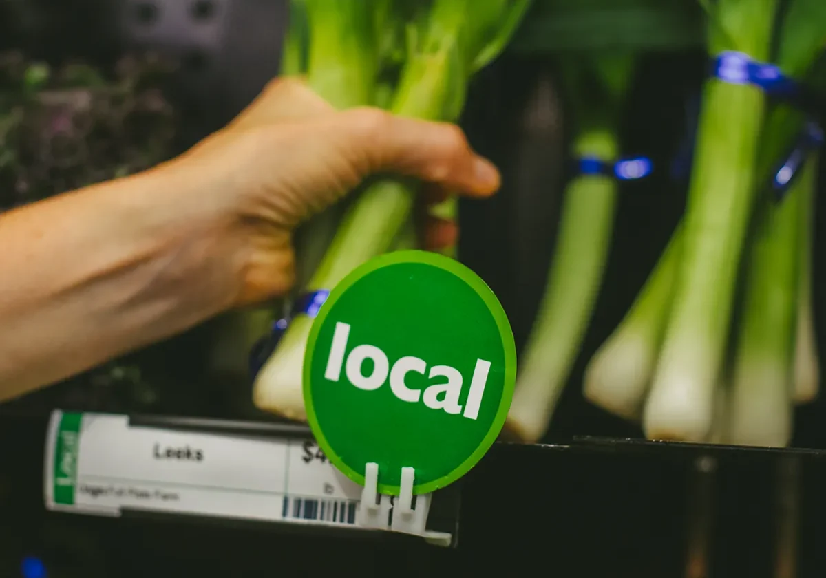 Hand reaching for leeks with a green local sign in front at a grocery store produce section.